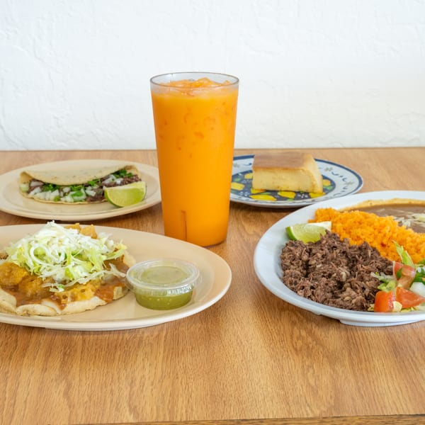 Mexican meal spread with tacos, shredded beef, rice, beans, salad, lime, cornbread, sauces, and an orange drink on a wooden table.