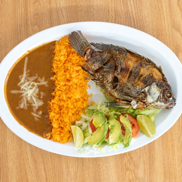 Plate of fried whole fish with orange rice, brown sauce, shredded cheese, lettuce, avocado, tomato, and lime wedges.