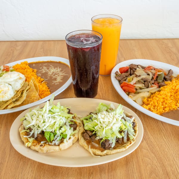 Mexican dishes with steak tacos, rice, beans, eggs, chips, salad, and two colorful drinks arranged on a wooden table.
