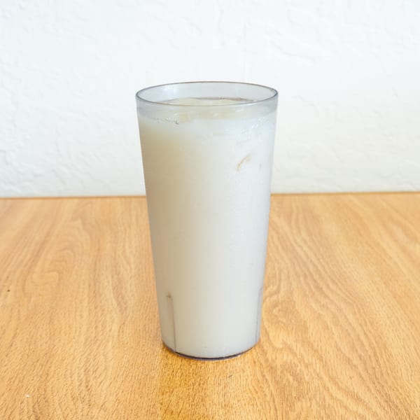 A tall glass of chilled, cloudy white beverage sits on a wooden table against a white wall.