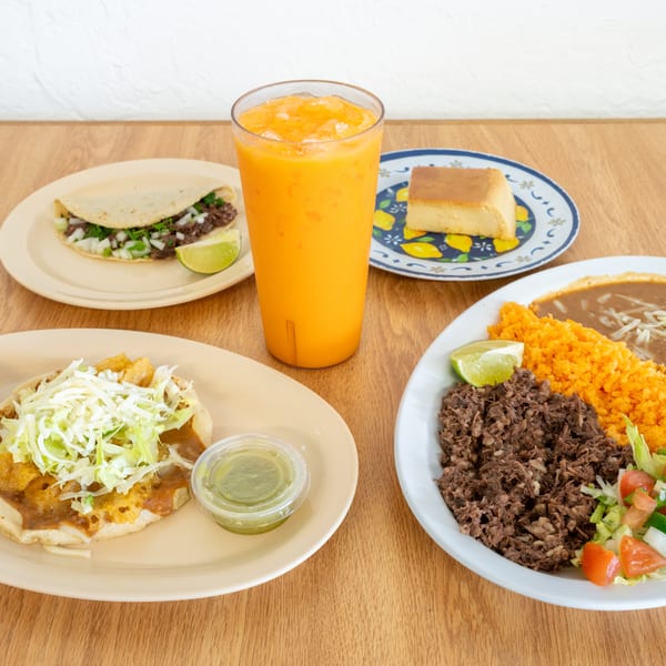 Mexican meal spread with tacos, shredded beef, rice, beans, cake, lime, salsa, and a glass of orange drink on a wooden table.